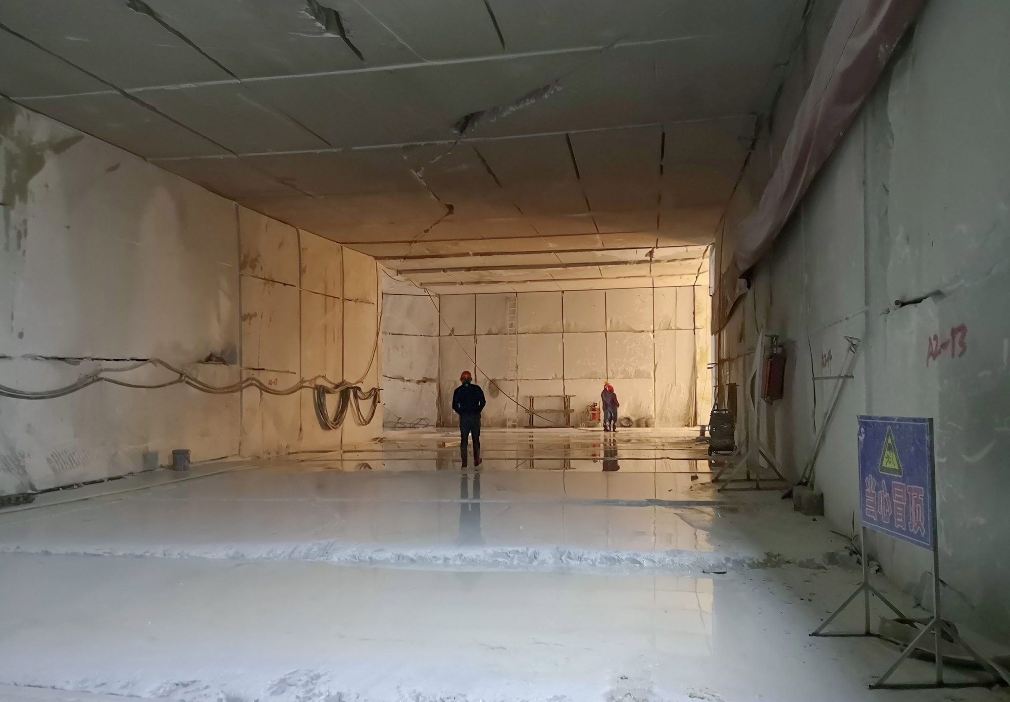 Inside a mining chamber: sawn white marble walls and ceiling, wet floor reflecting the light, two workers in red helmets at the far wall