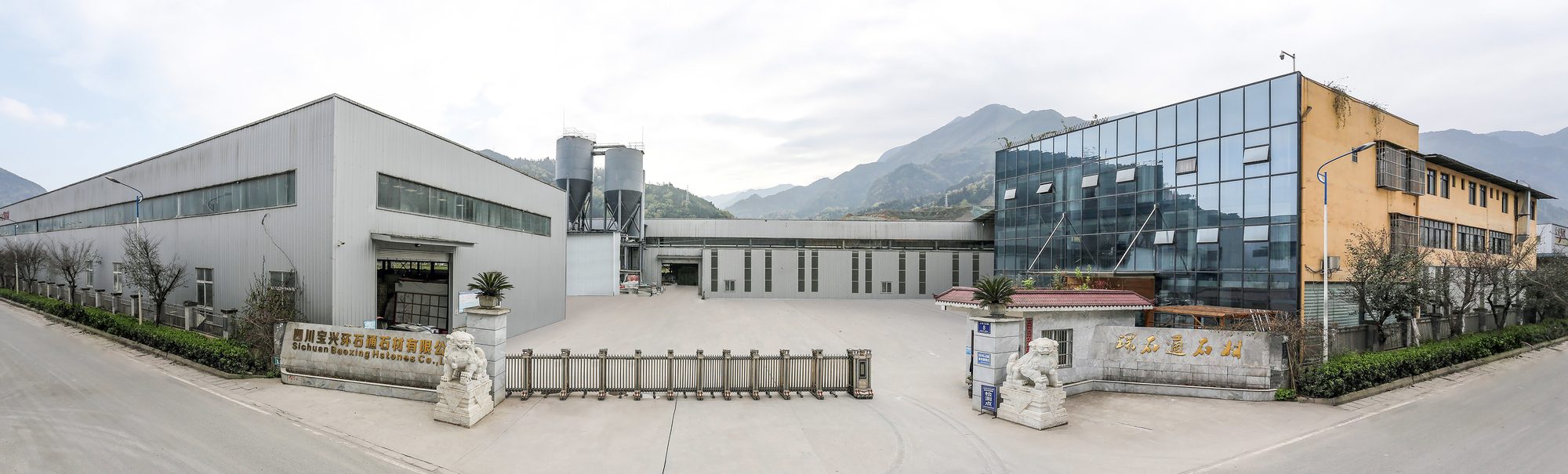 Panorama of the Huanshitong plant in Baoxing: steel-clad workshops, two silos, a glass fronted office block and the gate with two stone lions, mountains behind